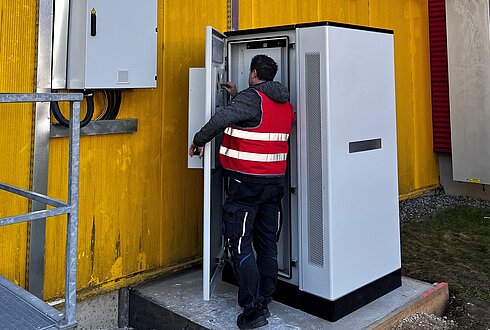 Person in Warnweste arbeitet an einem geöffneten Schaltschrank vor einer gelben Gebäudewand, daneben weitere Elektroinstallationen.