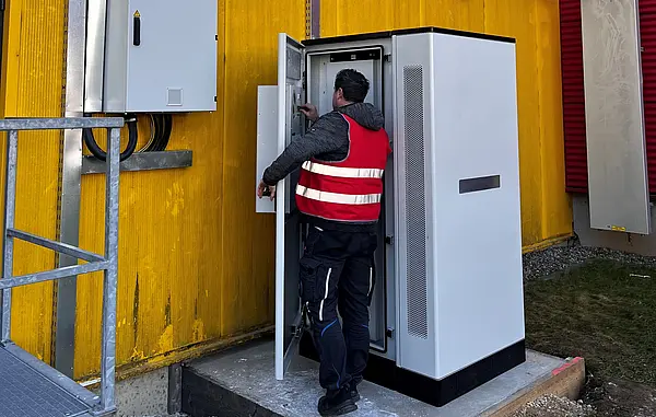 Person in Warnweste arbeitet an einem geöffneten Schaltschrank vor einer gelben Gebäudewand, daneben weitere Elektroinstallationen.