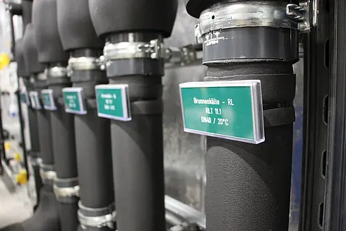 Mehrere isolierte Rohre mit Etiketten, die technische Angaben enthalten. Eine sichtbare Beschriftung lautet: „Brunnenkälte – RL, RLT 11.1, DN40 / 20°C“.