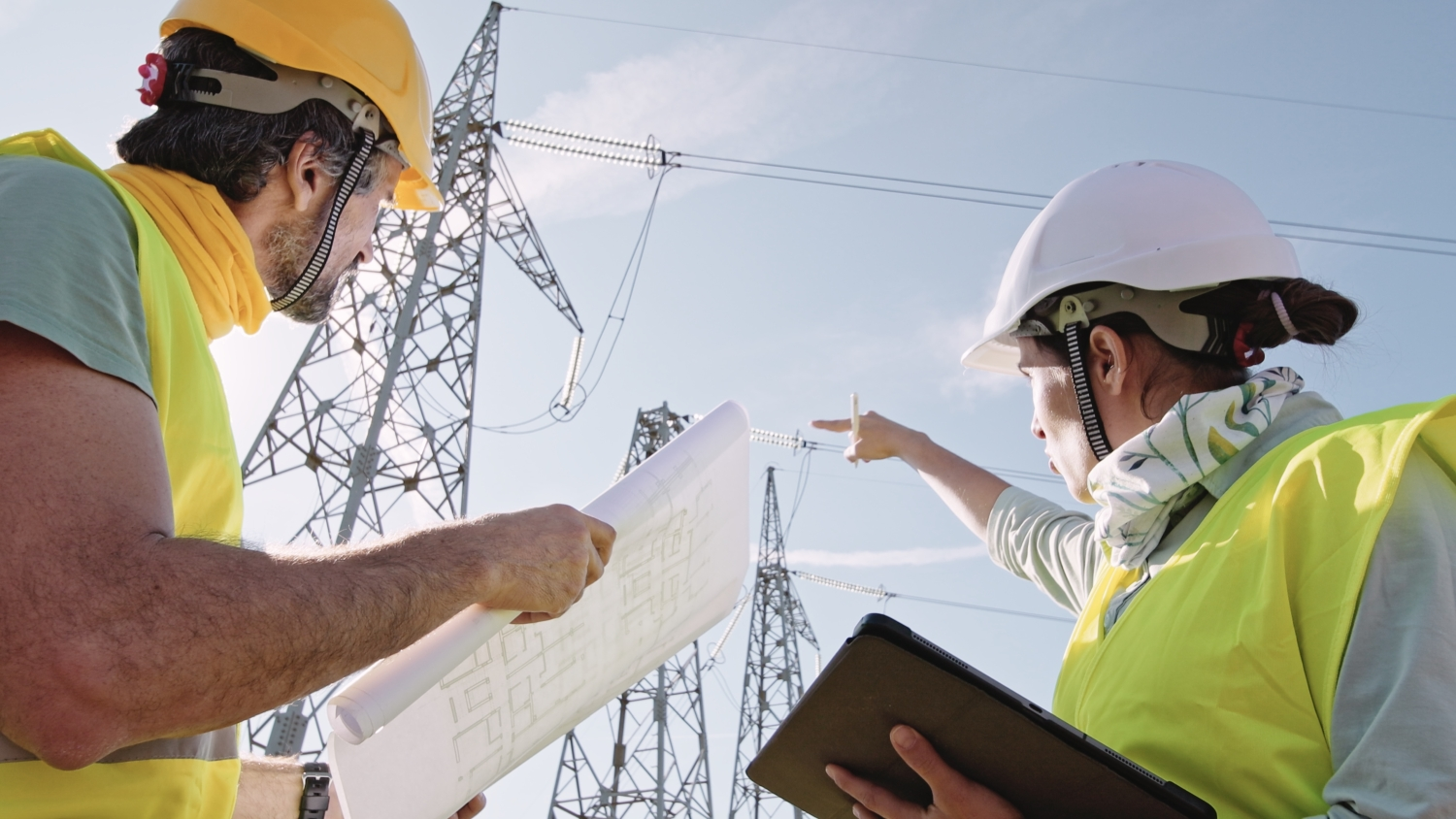 Zwei Personen stehen vor Strommasten. Die Frau hält Notizen und zeigt nach oben zu den Stromleitungen. Der Mann hält einen Plan und schaut nach oben. 