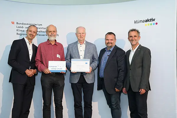 Menschen mit einer Urkunde in der Hand vor einer Fotowand. Sie lächeln in die Kamera.