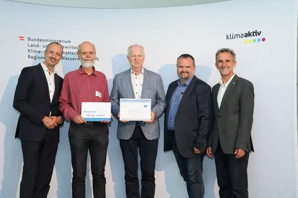 Menschen mit einer Urkunde in der Hand vor einer Fotowand. Sie lächeln in die Kamera.