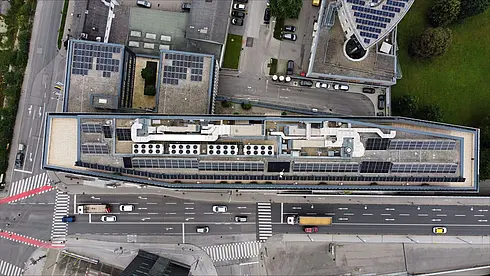 Luftaufnahme eines langen Gebäudes mit Solarpaneelen auf dem Dach, angrenzend an eine belebte Straße und städtische Umgebung.