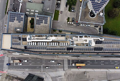 Luftaufnahme eines langen Gebäudes mit Solarpaneelen auf dem Dach, angrenzend an eine belebte Straße und städtische Umgebung.