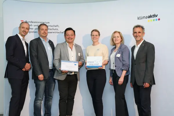 Menschen mit einer Urkunde in der Hand vor einer Fotowand. Sie lächeln in die Kamera.