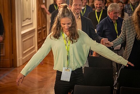 Renate Platzer mit geschlossenen Augen im großen Saal, im Hintergrund weitere Personen mit geschlossenen Augen