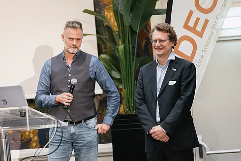 Zwei Männer vor einem Rednerpult und Banner mit der Aufschrift ‚DECO – Eine Stimme für Energie‘, einer spricht ins Mikrofon, der andere steht daneben.