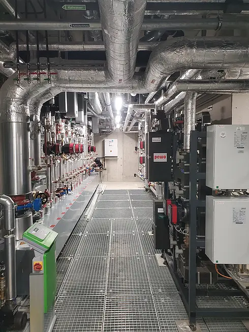 Technikraum mit isolierten Rohrleitungen entlang der Decke und Wände, mehreren Ventilen, Steuerungseinheiten und elektrischen Schaltkästen auf der rechten Seite. Der Boden besteht aus Metallgitter zur Wartung und Entwässerung.