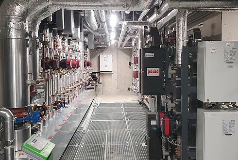 Technikraum mit isolierten Rohrleitungen entlang der Decke und Wände, mehreren Ventilen, Steuerungseinheiten und elektrischen Schaltkästen auf der rechten Seite. Der Boden besteht aus Metallgitter zur Wartung und Entwässerung.