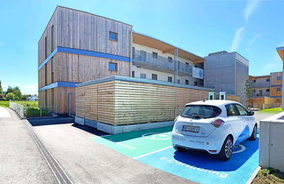 Ein Wohngebäude aus Holz, davor zwei Parkplätze für E-Autos. Ein Auto parkt darauf mit der Aufschrift "E-Carsharing"