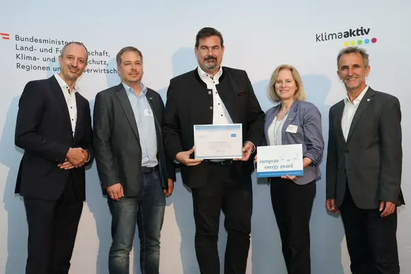 Menschen mit einer Urkunde in der Hand vor einer Fotowand. Sie lächeln in die Kamera.