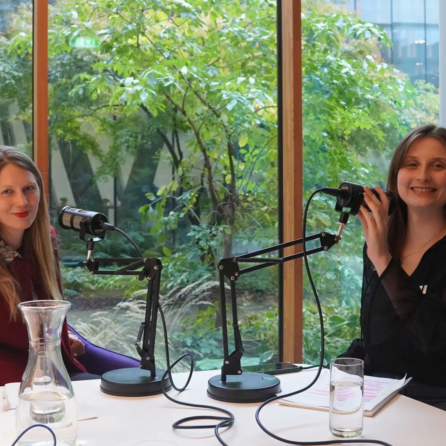 2 Frauen lächeln in die Kamera. Vor ihnen Mikrofone, hinter ihnen ein Garten.