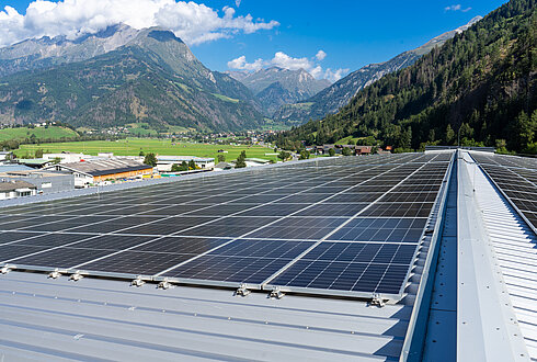 Große Solaranlage auf einem geneigten Metalldach, umgeben von grünen Feldern und Bergen unter klarem Himmel.