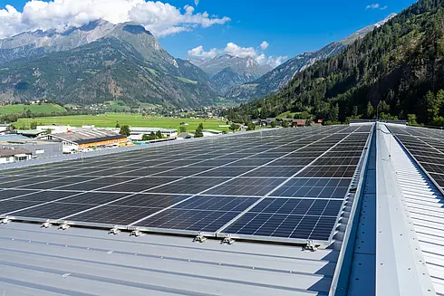 Große Solaranlage auf einem geneigten Metalldach, umgeben von grünen Feldern und Bergen unter klarem Himmel.