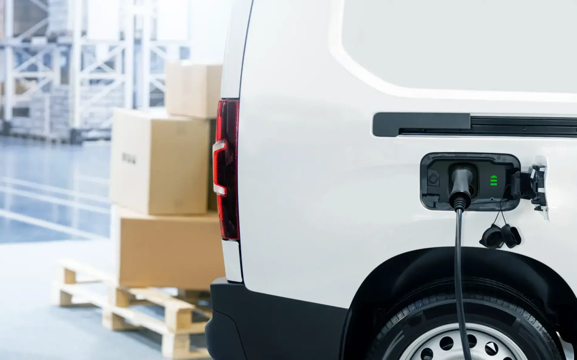 Der hintere Teil eines E-Lasters, der gerade mit einem Kabel geladen wird. Im Hintergrund Kartons und Paletten.