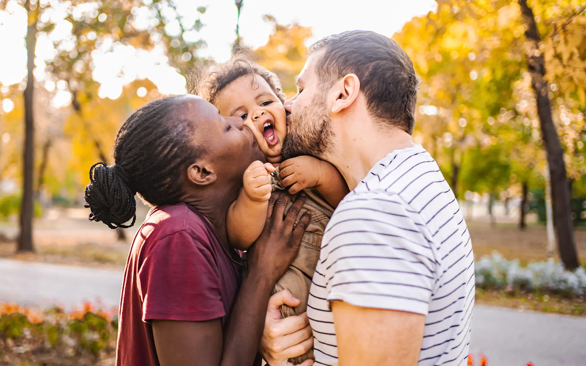 Fotografie einer kleinen Familie im Freien: Zwei Erwachsene halten ein Kleinkind in der Mitte und berühren mit ihren Gesichtern jeweils eine Wange des Kindes. Das Kind wird auf Brusthöhe getragen und schaut nach vorne, der Mund ist geöffnet. Alle drei Personen stehen nah beieinander und haben Körperkontakt. Die Szene spielt in einem Park mit unscharfem Hintergrund aus Bäumen in warmen Herbstfarben (gelb, orange und grün). Im Vordergrund sind zusätzlich Blumen und ein Weg erkennbar. Das Licht ist weich und warm, vermutlich spätnachmittags oder abends, und fällt seitlich auf die Personen. Die gesamte Bildstimmung wirkt hell und natürlich.