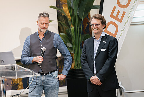 Zwei Männer vor einem Rednerpult und Banner mit der Aufschrift ‚DECO – Eine Stimme für Energie‘, einer spricht ins Mikrofon, der andere steht daneben.