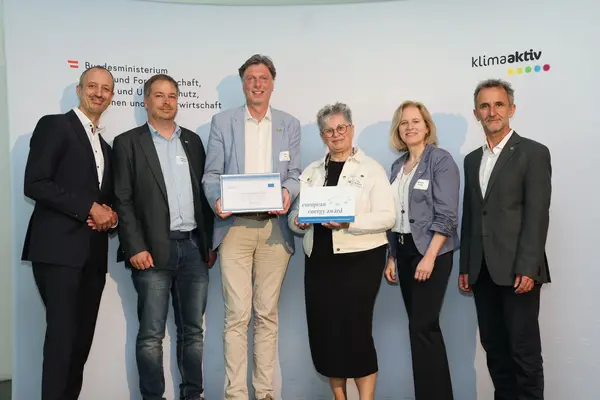 Menschen mit einer Urkunde in der Hand vor einer Fotowand. Sie lächeln in die Kamera.