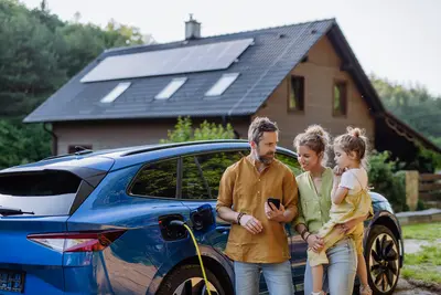 Ein Mann, eine Frau und ein Kind stehen vor ihrem E-Auto, das geladen wird. Im Hintergund ist ein Dach mit PV Panel zu sehen. 