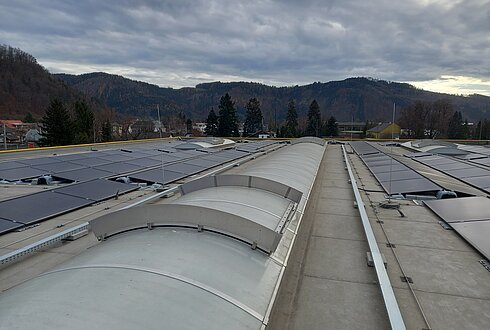 Flachdach mit installierten Solarmodulen und Lichtkuppeln, dahinter bewaldete Hügel unter teilweise bewölktem Himmel.