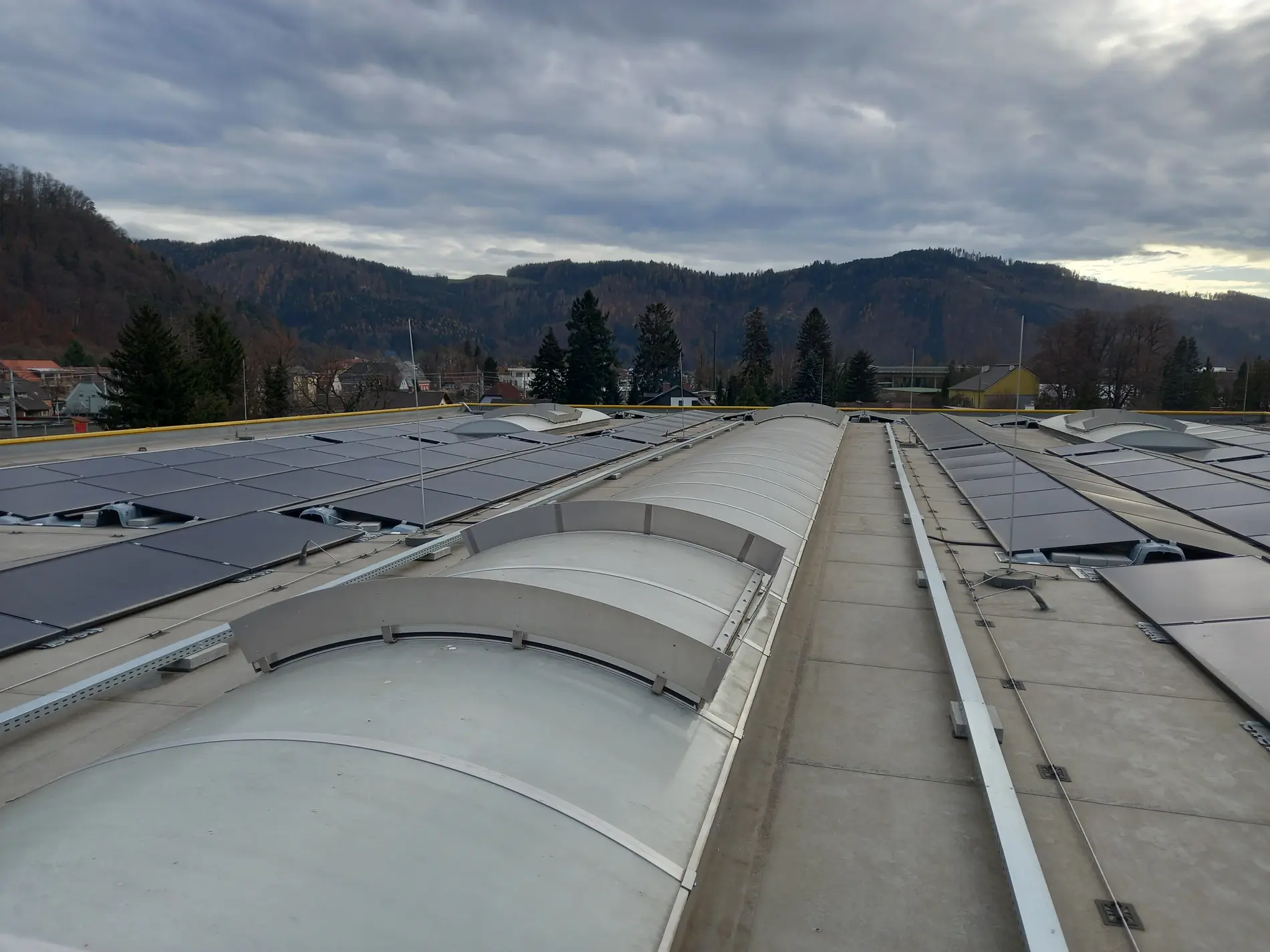 Flachdach mit installierten Solarmodulen und Lichtkuppeln, dahinter bewaldete Hügel unter teilweise bewölktem Himmel.