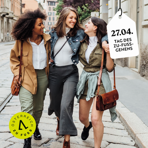 Drei Frauen gehen in einer Fußgängerzone sich gegenseitig umarmend und lachend. Im rechten Eck ein hängt ein digitales Schild mit der Aufschrift "27. 4. Tag des Zu-Fuß-Gehens"
