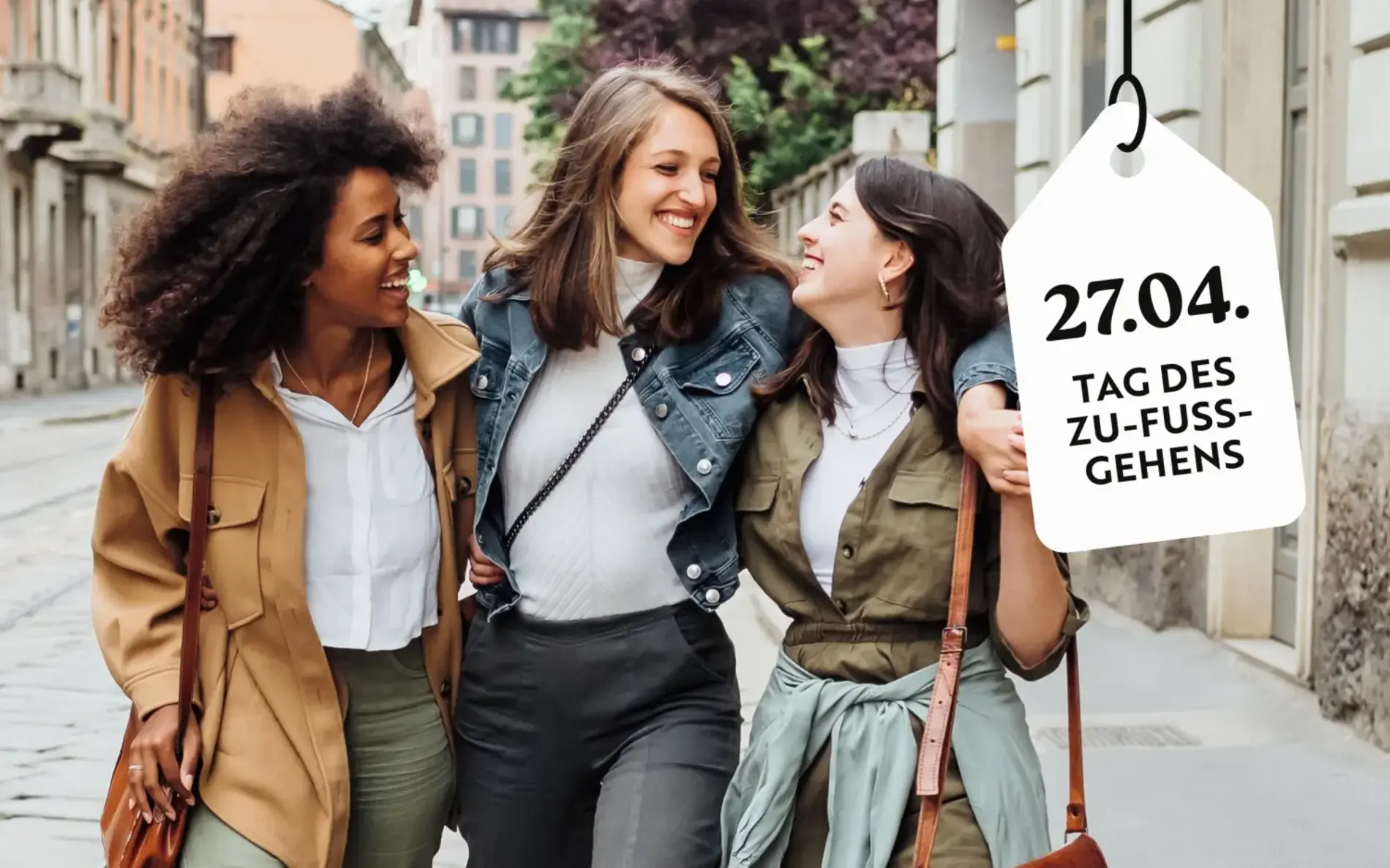 Drei Frauen gehen in einer Fußgängerzone sich gegenseitig umarmend und lachend. Im rechten Eck ein hängt ein digitales Schild mit der Aufschrift "27. 4. Tag des Zu-Fuß-Gehens"