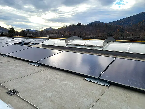 Flachdach mit installierten Solarmodulen und Lichtkuppeln, dahinter bewaldete Hügel unter teilweise bewölktem Himmel