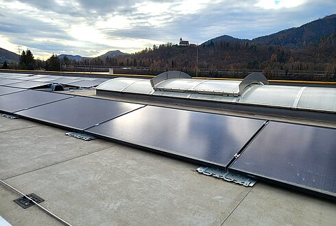 Flachdach mit installierten Solarmodulen und Lichtkuppeln, dahinter bewaldete Hügel unter teilweise bewölktem Himmel