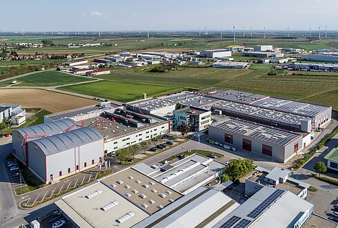 Luftaufnahme eines Industriegebiets mit mehreren Hallen, Solaranlagen auf den Dächern und Windkraftanlagen im Hintergrund.