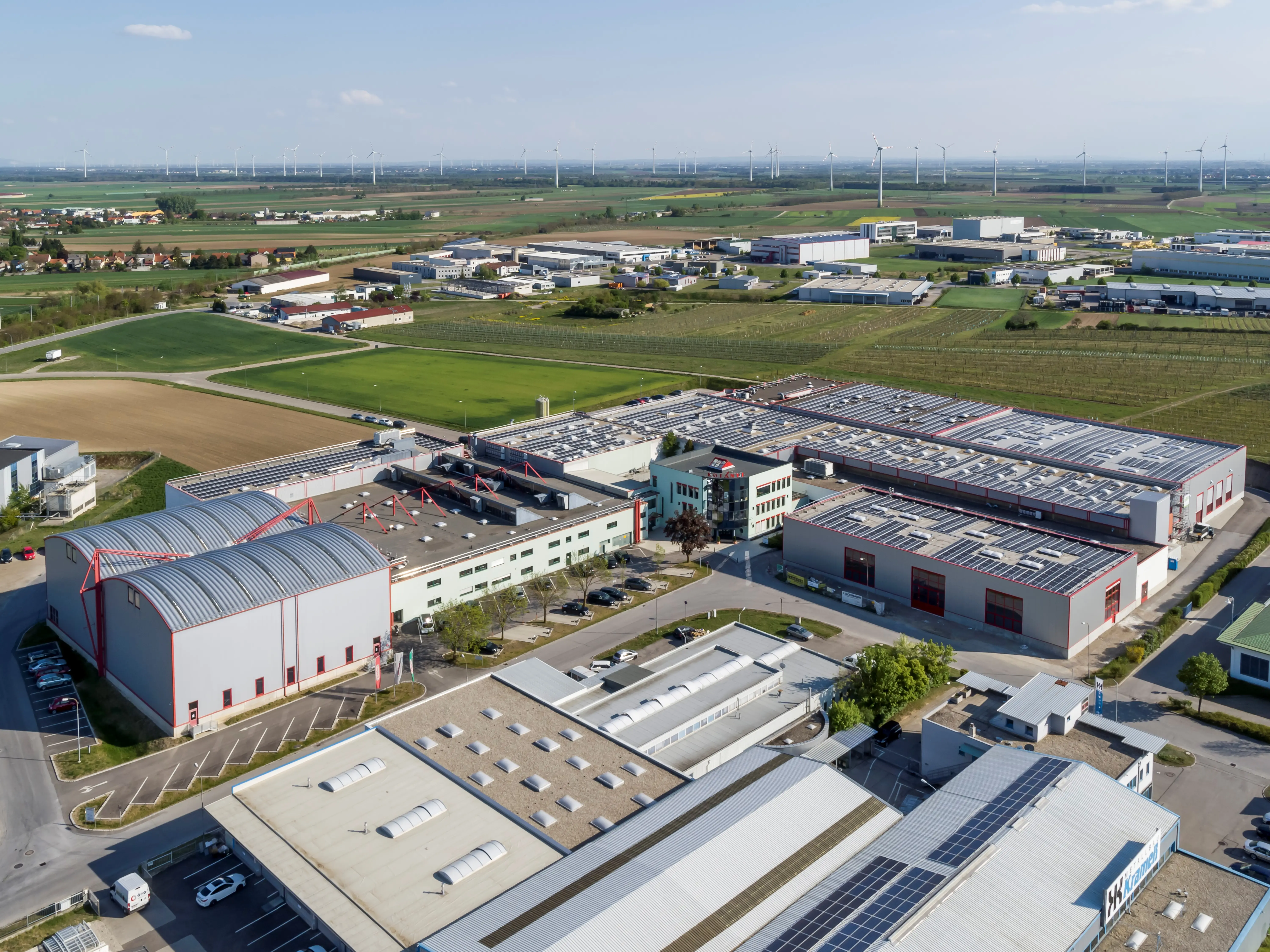 Luftaufnahme eines Industriegebiets mit mehreren Hallen, Solaranlagen auf den Dächern und Windkraftanlagen im Hintergrund.